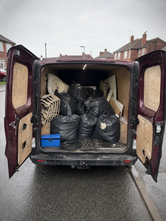 A burgundy panel van with rear doors open, loaded with black refuse sacks, cardboard and broken furniture