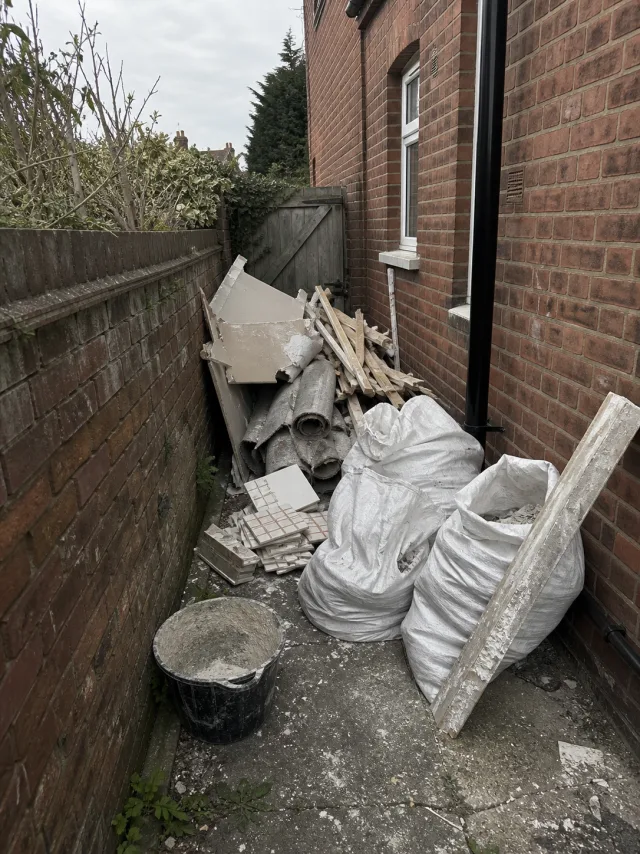 Broken plasterboard, stud timber, tiles and filled rubble sacks piled along a side return beside a red brick house