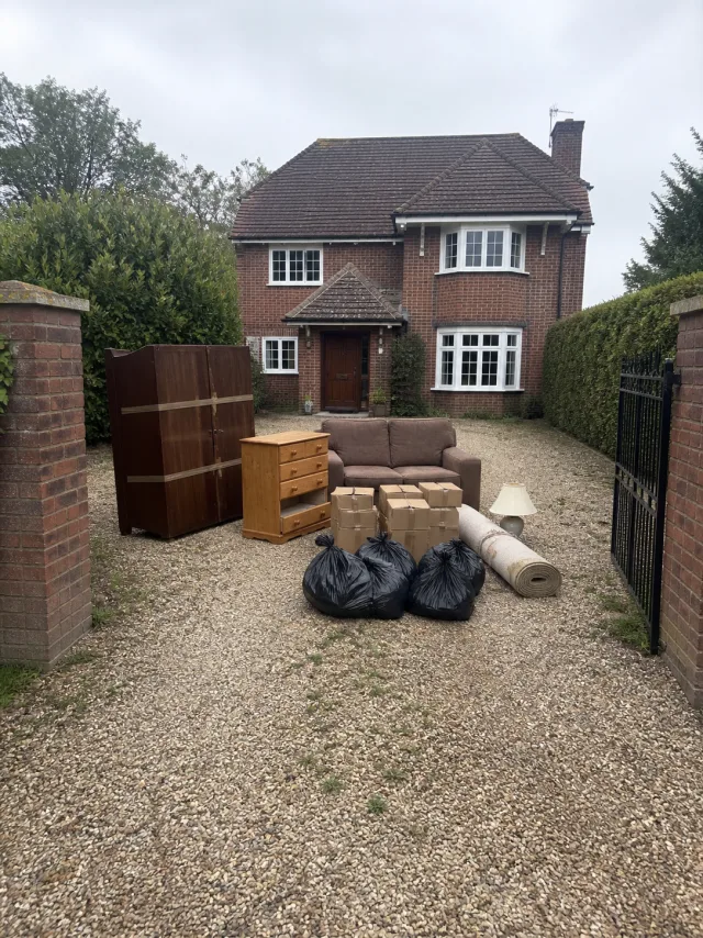 A wardrobe, chest of drawers, sofa, boxes, rolled carpet and refuse sacks set out on a gravel drive in front of a large detached house