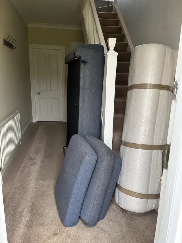 A sofa stood on end with its cushions removed and a wrapped mattress leaning against the stairs in a house hallway