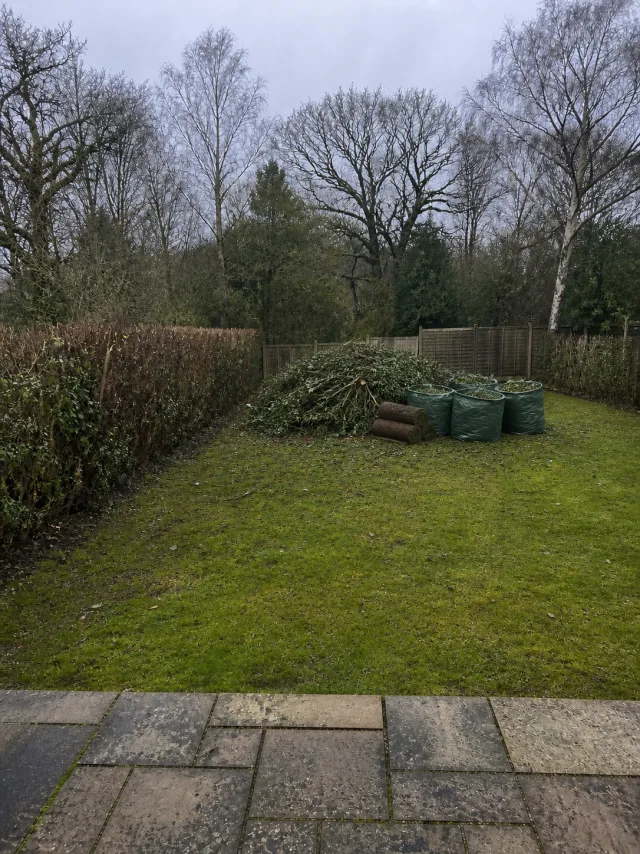 A heap of cut branches, filled green garden waste bags and rolled turf on the lawn of a large garden backing onto woodland