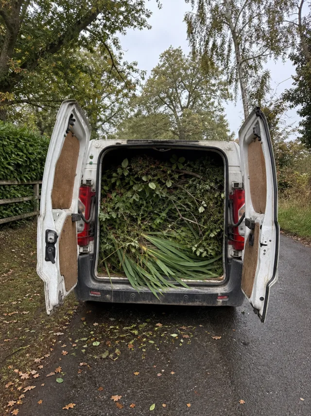 A white panel van with rear doors open on a country lane, filled to the roof with mixed green waste