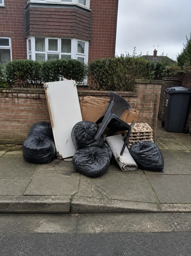 A pile of black refuse sacks, a broken cabinet door, a garden chair and rolled carpet stacked on a suburban pavement